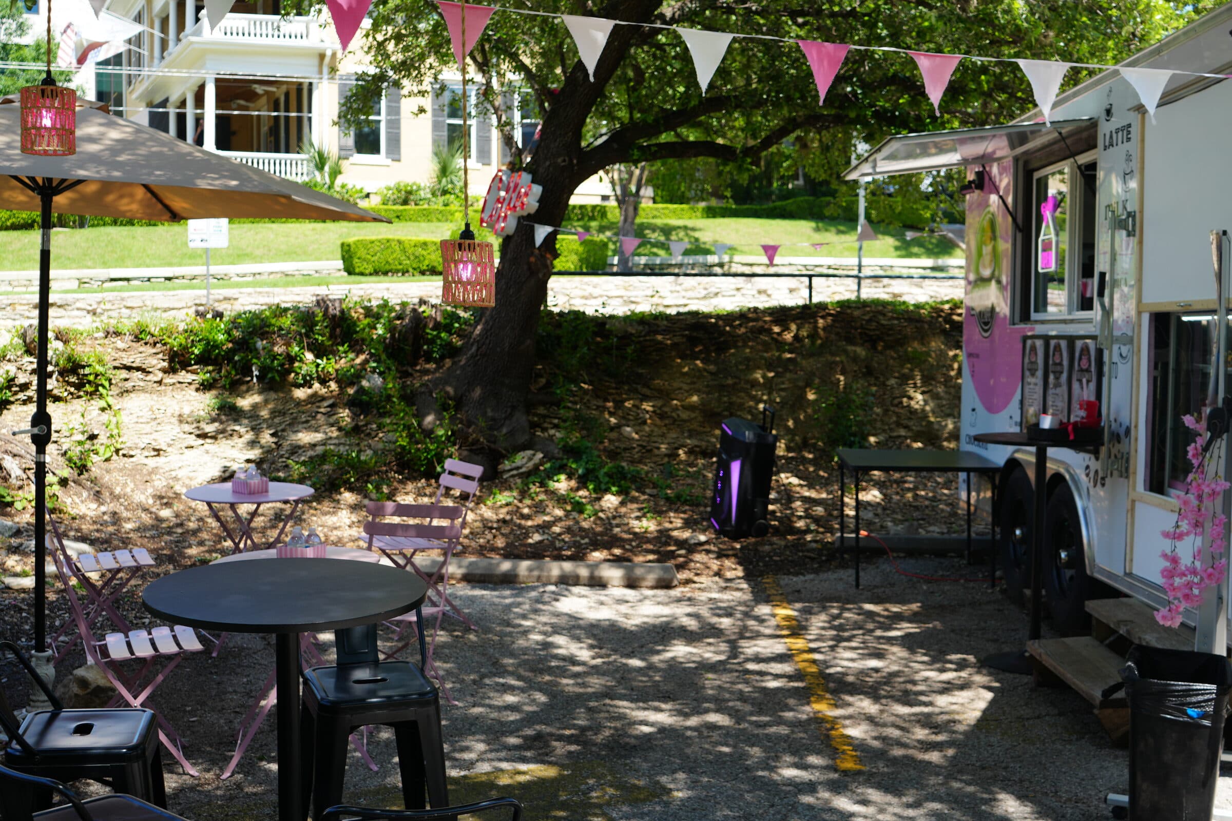 Coffee truck patio on the ANW lot — pink umbrella, café tables, and bunting under a tree